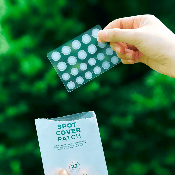 Ruka drží hárok s náplasťami Spot Cover Patches, ktoré sú priehľadné a zaoblené. V pozadí je rozmazané zelené prostredie. Balenie ukazuje časť balenia s textom "SPOT COVER PATCH" a informáciou, že produkt obsahuje 22 náplastí, ktoré pomáhajú liečiť malé oblasti pokožky.