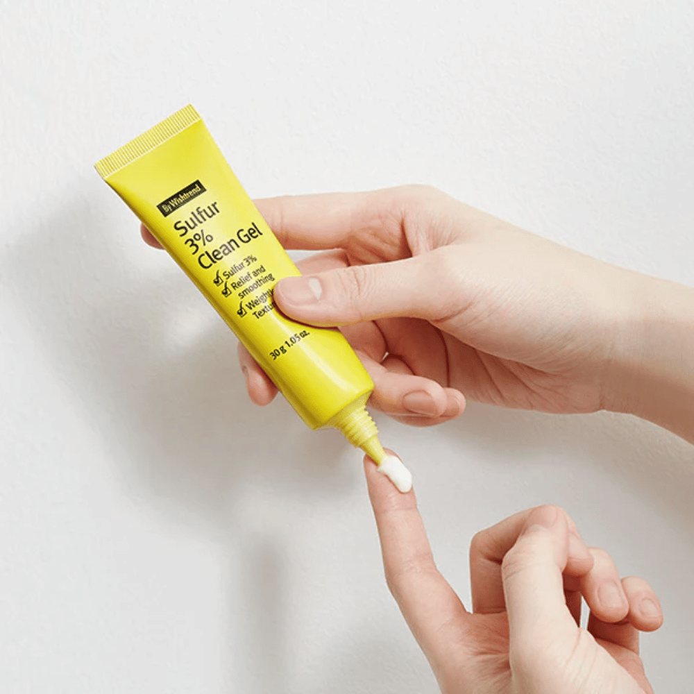 Detailný záber na ruky, ktoré aplikujú By Wishtrend Sulfur 3% Clean Gel, žltú tubu s bielym gélom, na bielom pozadí.