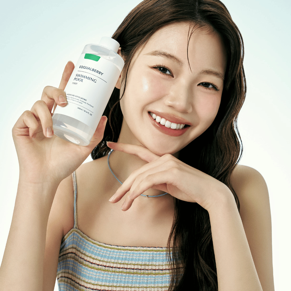 Usmievajúca sa žena so žiarivou pleťou drží Equalberry Swimming Pool Toner. Svieži a prirodzený vzhľad s ľahkým mejkapom a letným outfitom v modrých tónoch.