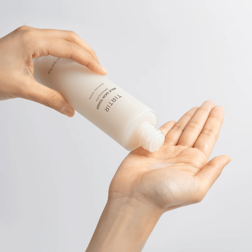 Obrázok ukazuje osobu, ktorá vylieva mliečnu tekutinu z fľaše označenej "TIRTIR Milk Skin" na dlaň. Produkt sa javí ako tonikum na pleť alebo hydratačné mlieko.
