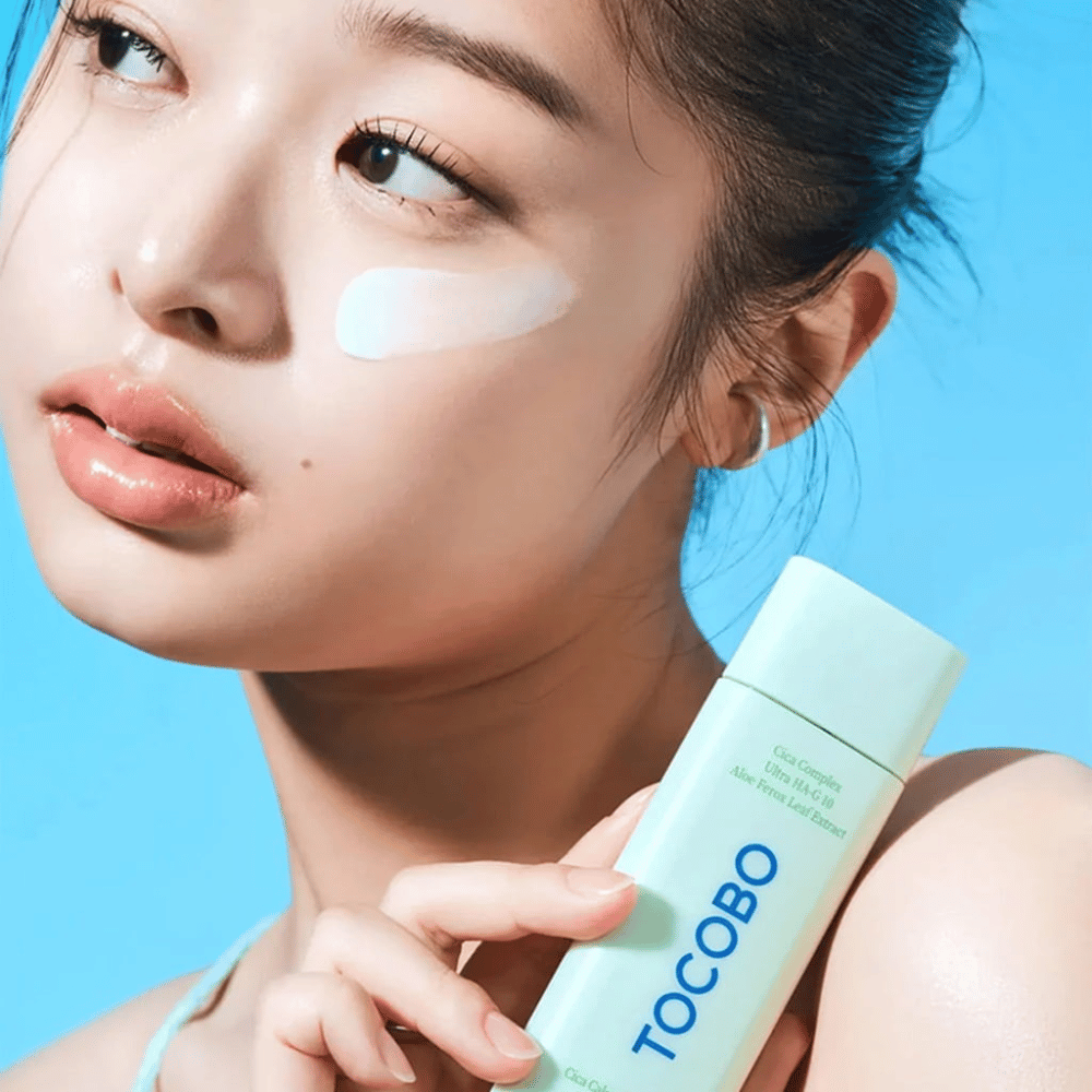 Modelka s opaľovacím krémom pod okom drží TOCOBO Cica Calming Sun Serum v mätovo zelenom balení na modrom pozadí.