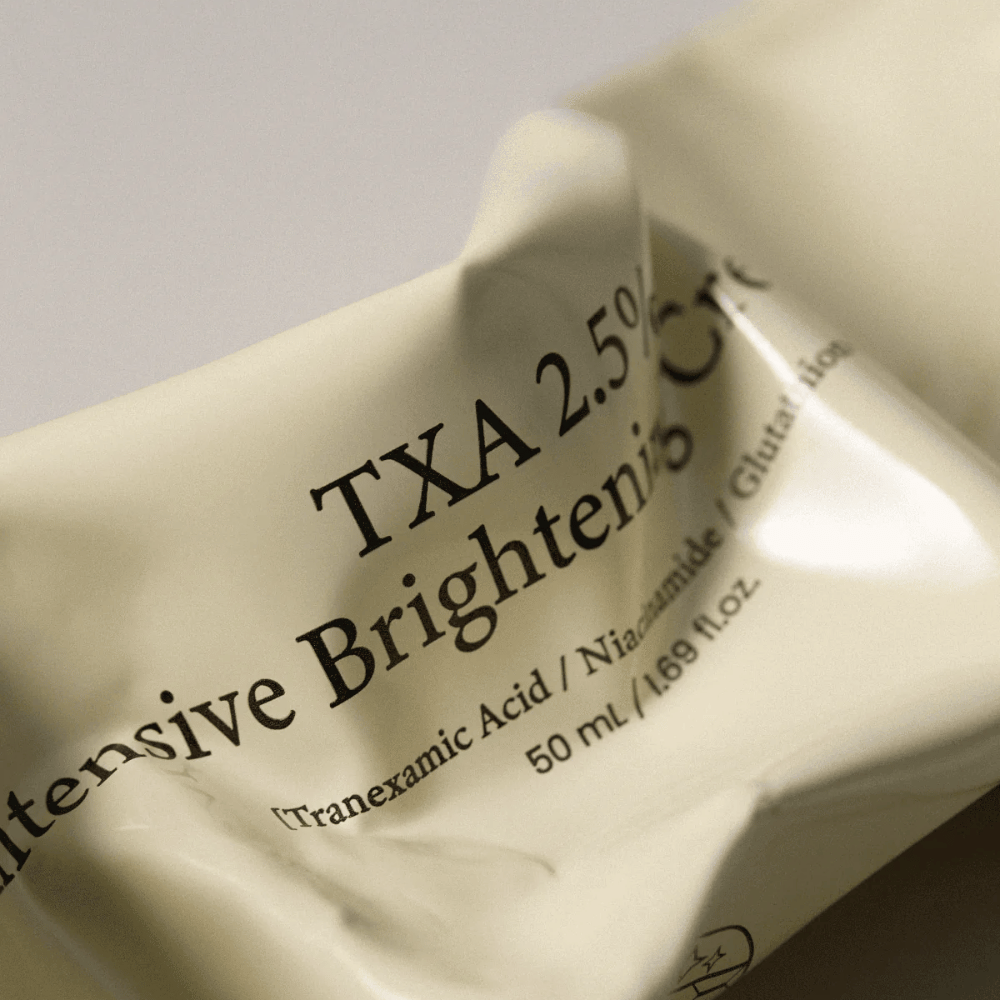Detailný záber na AXIS - Y TXA 2.5% Intensive Brightening Cream, kde je v centre pozornosti jemný a minimalistický dizajn tuby. Obrázok zvýrazňuje názov produktu, Tranexamic Acid a Niacinamide, ako aj elegantné béžové balenie.