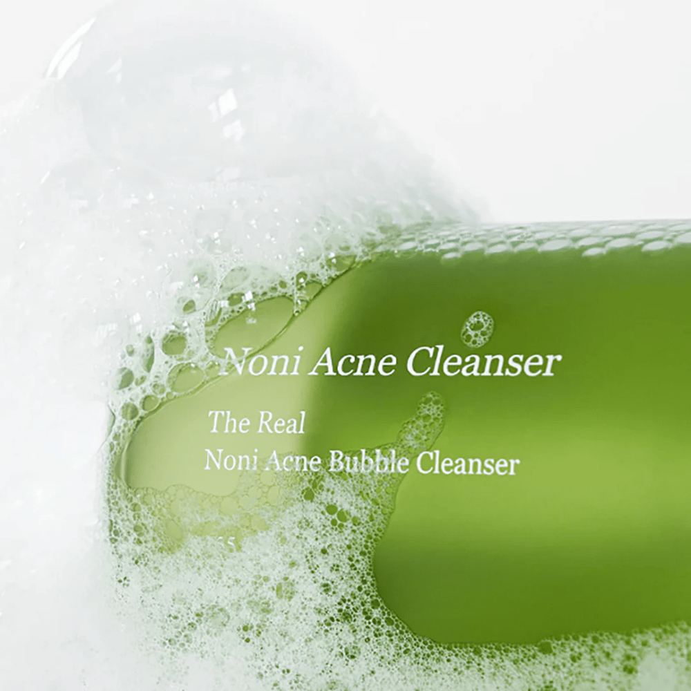 Fľaša s The Real Noni Acne Bubble Cleanser je v centre pozornosti a je obklopená bohatou penou. Svieža zelená farba signalizuje prírodné zložky, ideálne na čistenie a boj proti akné. Ideálny produkt pre čistú a jasnú pleť.