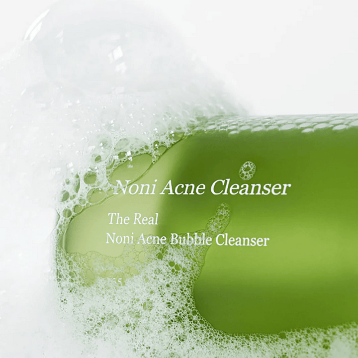 Fľaša s The Real Noni Acne Bubble Cleanser je v centre pozornosti a je obklopená bohatou penou. Svieža zelená farba signalizuje prírodné zložky, ideálne na čistenie a boj proti akné. Ideálny produkt pre čistú a jasnú pleť.