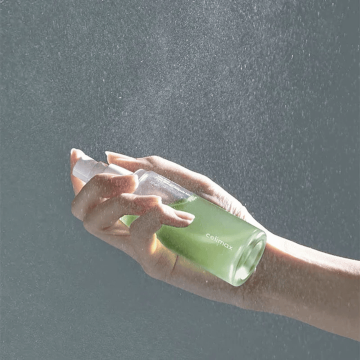 Ruka drží sprejovú fľašu Celimax Noni Mist a aplikuje produkt v jemnom, rovnomernom spreji na pokožku. Fľaša je zelená a produkt je propagovaný ako "The Real Noni Energy Ampule Mist" na dodanie energie a hydratácie pokožke.