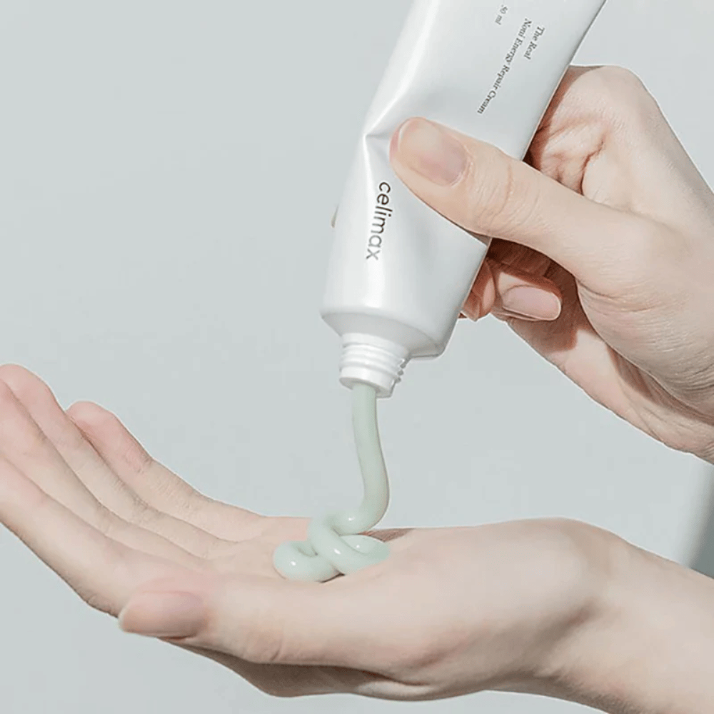 Ruka drží bielu tubu s Celimax Noni Energy Repair Cream a krém je vytlačený v rovnom prúde na dlaň. Krém je svetlozelený a je propagovaný na výživu a obnovu pokožky.