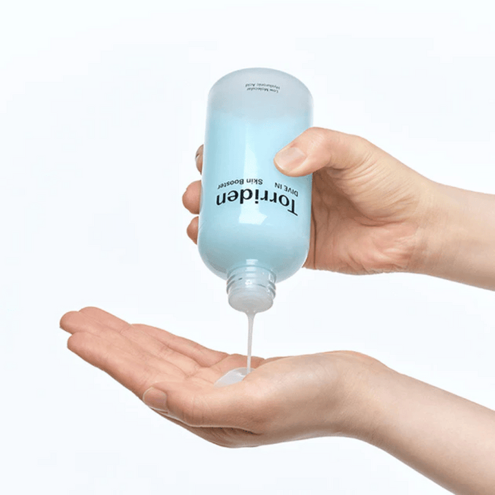 Ruky držiace fľašu s produktom starostlivosti o pleť a dávkujúce číru tekutinu do dlane. Fľaša je označená "Torriden - DIVE-IN Low Molecule Hyaluronic Acid Skin Booster".