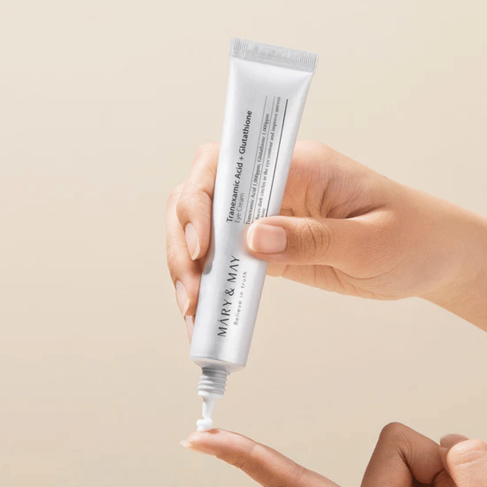 Ruka drží tubu MARY & MAY "Tranexamic Acid + Glutathione Eye Cream". Tuba je biela a dizajn je čistý a jednoduchý, čo zdôrazňuje upokojujúce a zosvetľujúce vlastnosti produktu pre oblasť očí. Produkt sa aplikuje jemne, s dôrazom na presnosť a efektívnosť.