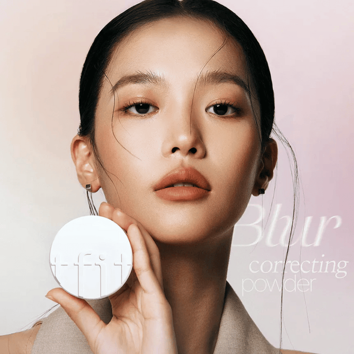 Žena s bezchybným make-upom drží okrúhlu bielu dózu s logom TFIT, s textom "Blur Correcting Powder" v pozadí.