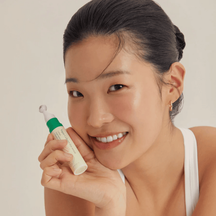 Žena so zviazanými vlasmi a zlatými náušnicami drží AXIS-Y Vegan Collagen Eye Serum 10ml blízko tváre, zatiaľ čo sa jemne usmieva. Sérum je navrhnuté na hydratáciu a redukciu vrások okolo očí, špeciálne vyvinuté pre citlivú pokožku.