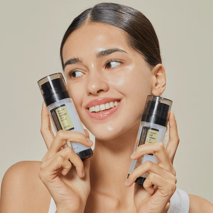 Usmievajúca sa žena s lesklou pokožkou drží dve fľaše COSRX Advanced Snail Radiance Dual Essence 80 ml blízko tváre na svetlom pozadí.