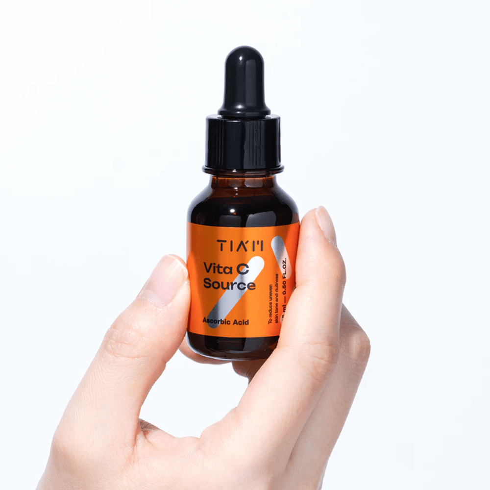 Ruka drží TIA'M Vita C Source, sérum s vitamínom C v hnedej sklenenej fľaši s pipetou. Produkt je navrhnutý na vyrovnanie tónu pleti a dodanie žiarivosti.