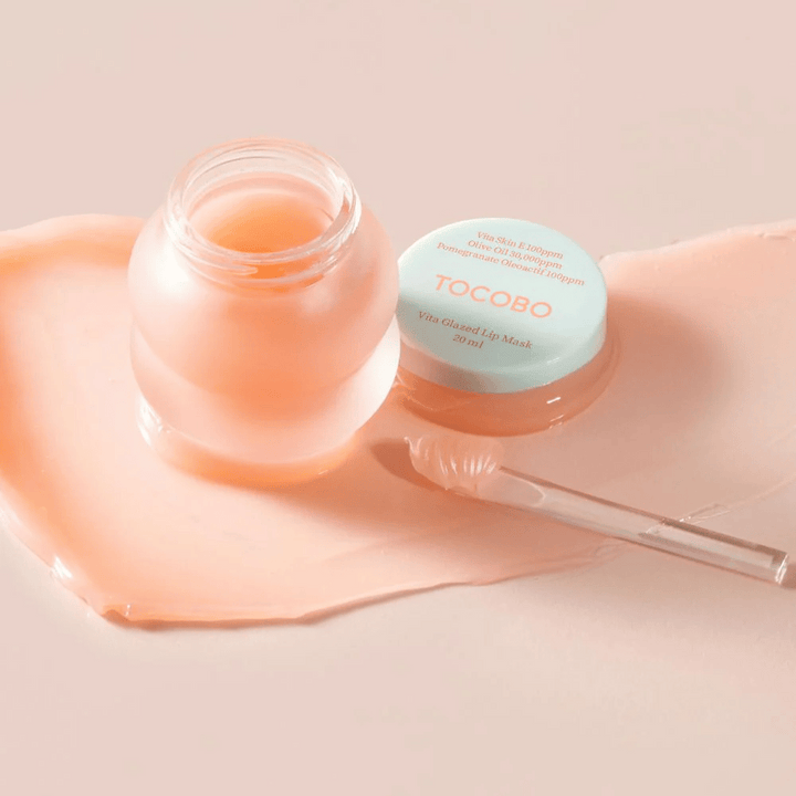 Zobrazuje sa nádoba s "Vita Glazed Lip Mask" od Tocobo. Nádoba je priehľadná so svetlo tyrkysovým viečkom a obsahuje lesklú, ružovú masku na pery. Malá špachtľa leží vedľa nádoby a časť masky je rozotretá na povrchu. Produkt je navrhnutý na hydratáciu a starostlivosť o pery.