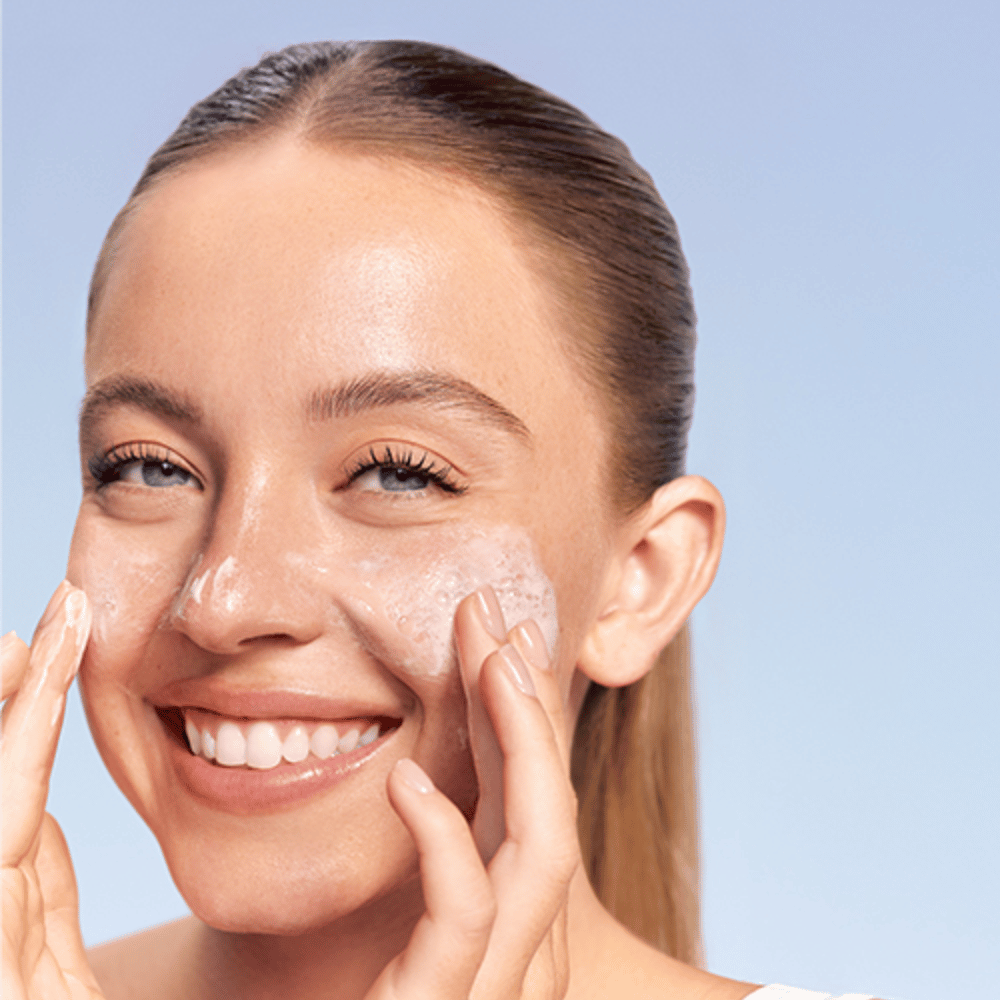 Usmievajúca sa žena aplikuje LANEIGE Water Bank Gentle Gel Cleanser v penovej forme na líca, na modrom pozadí.