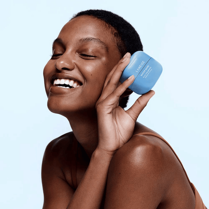 Usmievavá žena drží modrú nádobu LANEIGE Water Bank Blue Hyaluronic Cream pri líci, so svetlomodrým pozadím.