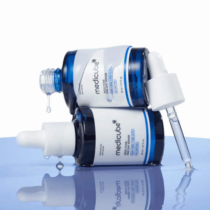 Dve modré fľaše Medicube Zero Pore One Day Serum ležiace oproti sebe, s pipetami a kvapkami séra na reflexnej modrej ploche.