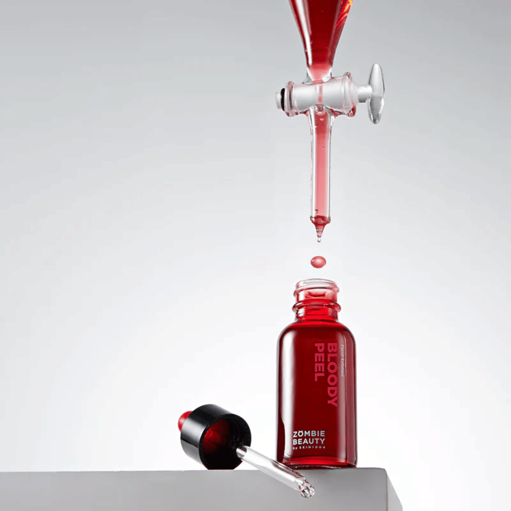 Fľaša Zombie Beauty Bloody Peel v červenom skle, s pipetou vedľa. Kvapka červenej tekutiny padá z pipety nad fľašou a smeruje do nej. Pozadie je svetlosivé a zvýrazňuje dramatickú farbu produktu.