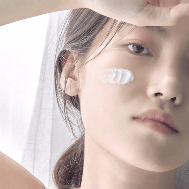 Detailný záber na mladú ženu s kvapkou krému na líci a rukou zdvihnutou nad čelo proti jemnému pozadiu.