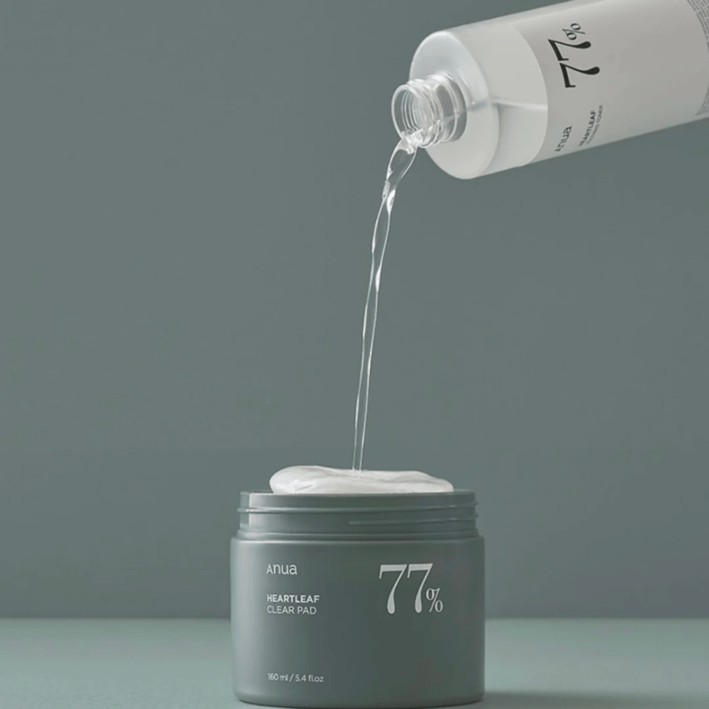 Priehľadná fľaša nalieva tekutinu do šedej nádoby označenej "anua HEARTLEAF 77% Clear Pad", ktorá obsahuje stoh bielych vankúšikov na starostlivosť o pleť. Pozadie je jednoduché a jednofarebné v jemnom šedom tóne.