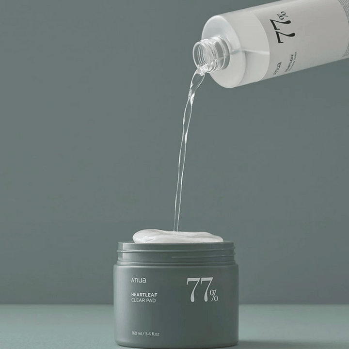 Priehľadná fľaša nalieva tekutinu do šedej nádoby označenej "anua HEARTLEAF 77% Clear Pad", ktorá obsahuje stoh bielych vankúšikov na starostlivosť o pleť. Pozadie je jednoduché a jednofarebné v jemnom šedom tóne.
