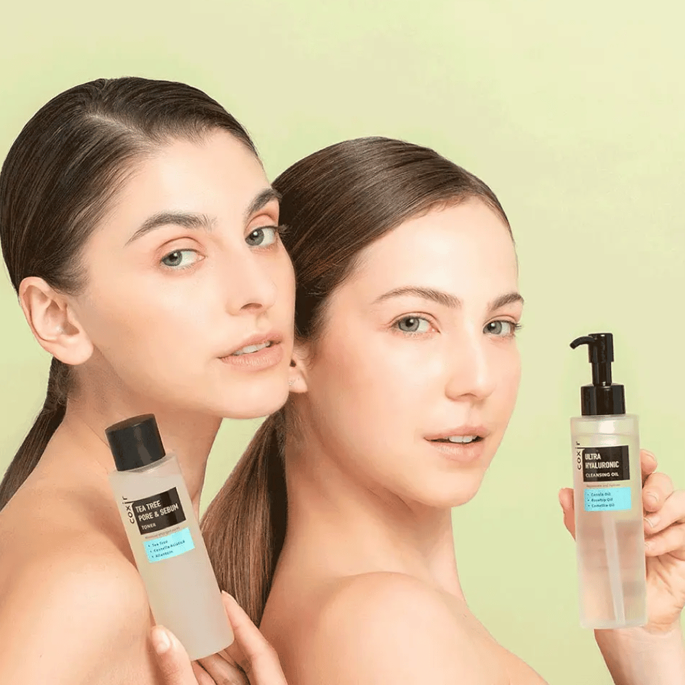 Dve ženy s odhalenými ramenami pózujú s kozmetickými produktmi; jedna drží fľašu označenú "TEA TREE PORE & SEBUM", a druhá pumpovaciu fľašu označenú "ULTRA HYALURONIC CLEANSING OIL". Stoja pred svetlozeleným pozadím.