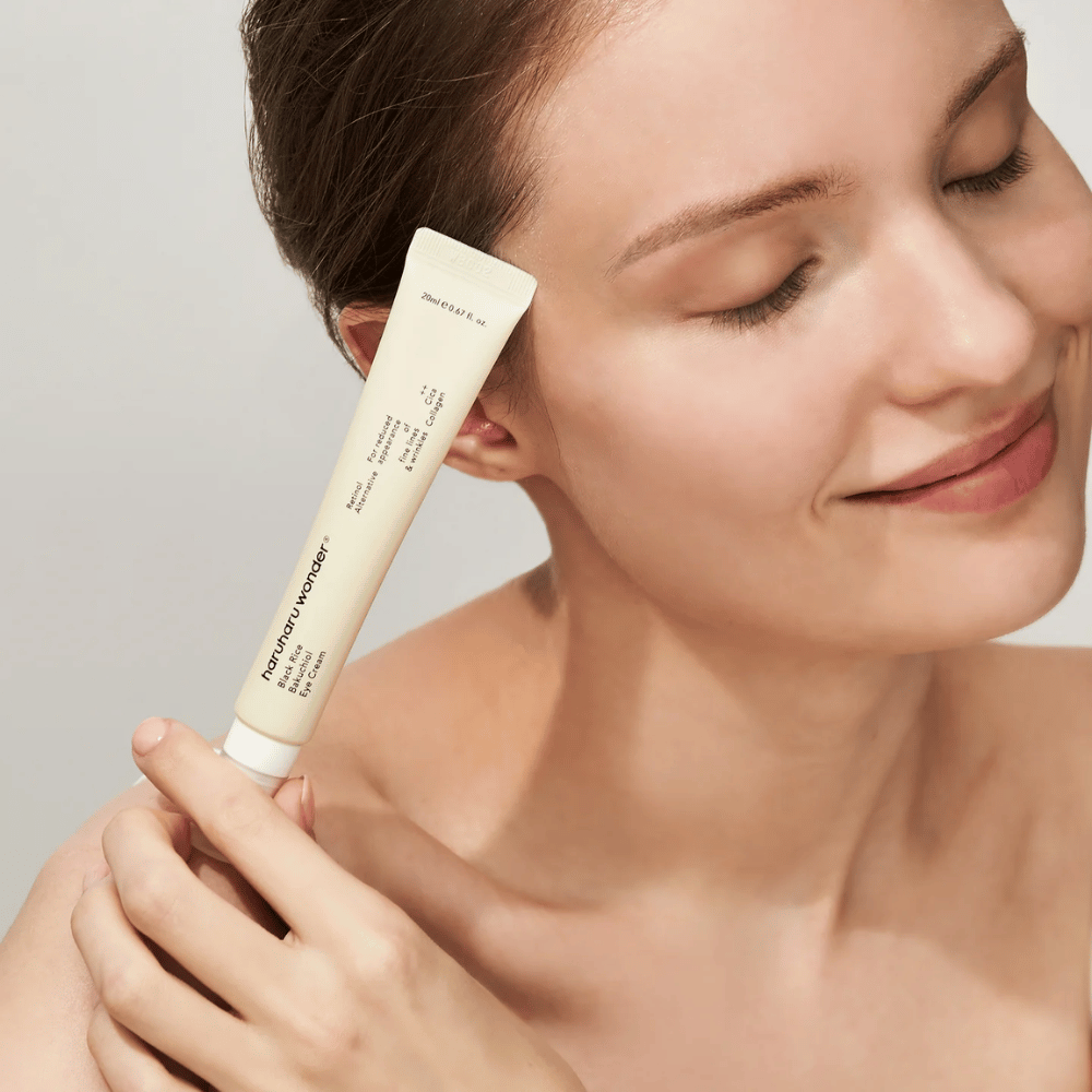Usmievajúca sa osoba so zatvorenými očami drží tubu Haruharu Wonder Black Rice Bakuchiol Eye Cream pri spánkoch.