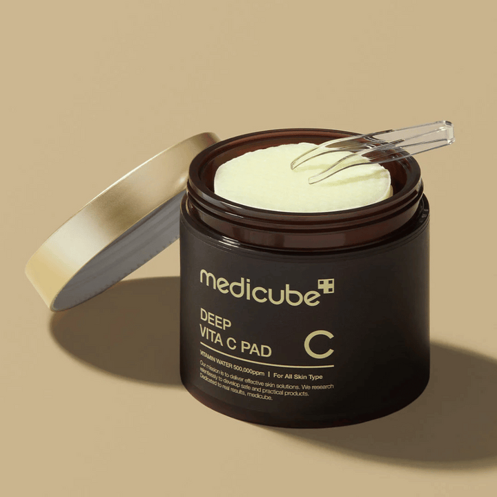 Pohár s medicube Deep Vita C Pad. Pohár je tmavohnedý so zlatým textom a bielou, inverznou etiketou s informáciou o vitamíne C. Vnútro viečka je zlaté a priehľadná pinzeta leží na svetložltých bavlnených tampónoch vo vnútri. Pozadie je jednofarebné béžové.