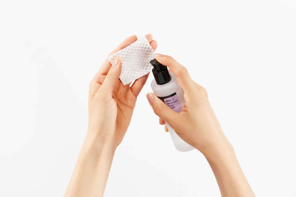 Ruka držiaca fľašu čistiaceho prostriedku s COSRX AHA 7 Whitehead Power Liquid 100ml na liečbu akné na bielom pozadí.