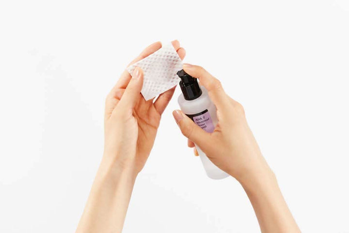 Ruka držiaca fľašu čistiaceho prostriedku s COSRX AHA 7 Whitehead Power Liquid 100ml na liečbu akné na bielom pozadí.