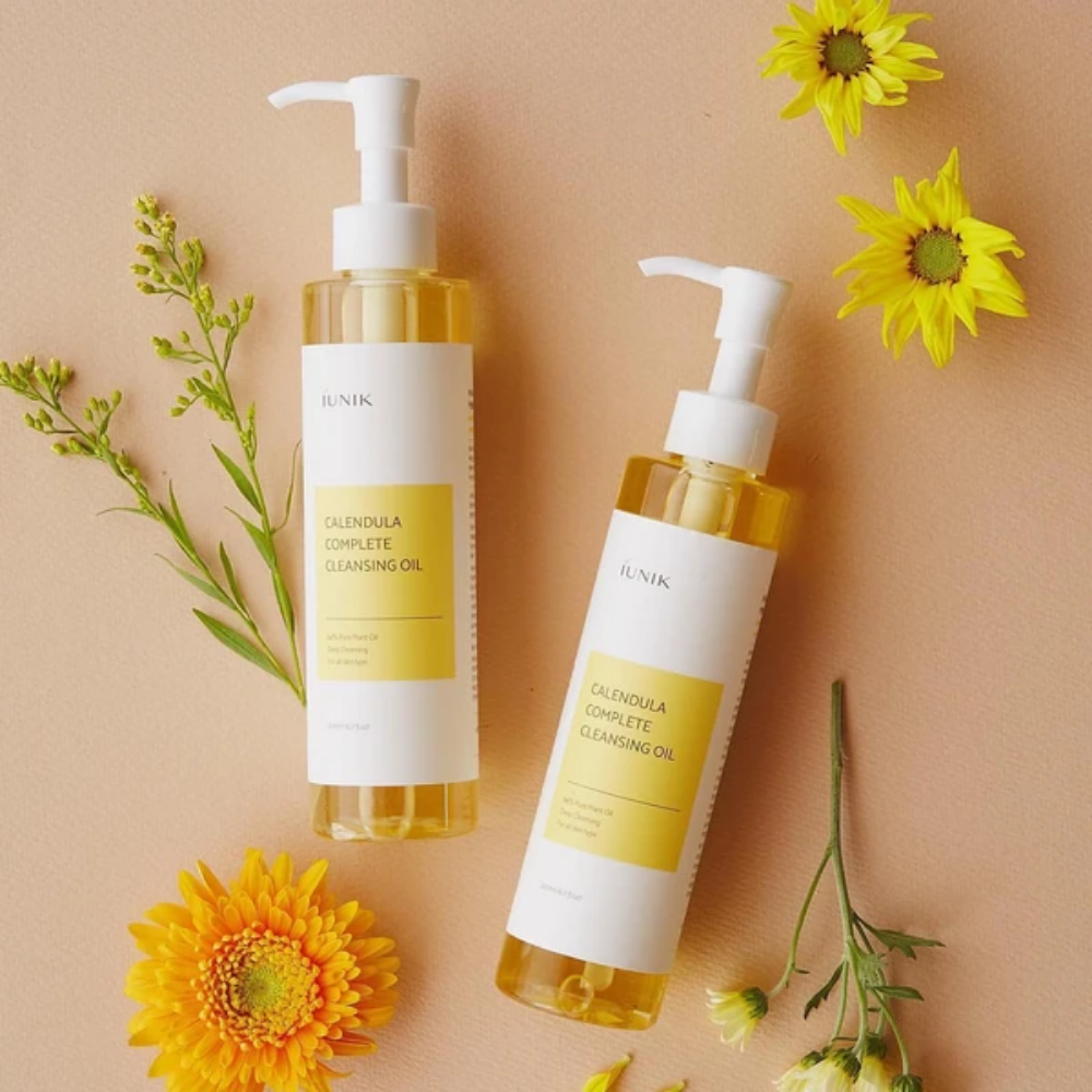 Dve fľaše iUNIK Calendula Complete Cleansing Oil 200ml so žltými kvetmi na béžovom pozadí, naplnené rastlinným olejom.