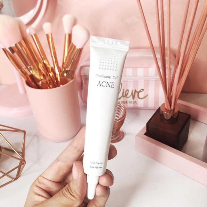 Osoba držiaca tubu Pyunkang Yul Acne Spot Cream 15 ml pred ružovým stolom.