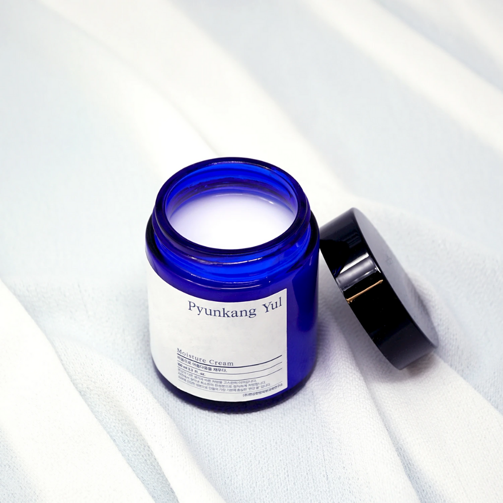 Modrý pohár s viečkom, ktorý je na bielej tkanine a ukazuje hydratačné vlastnosti Pyunkang Yul's Moisture Cream 100ml.