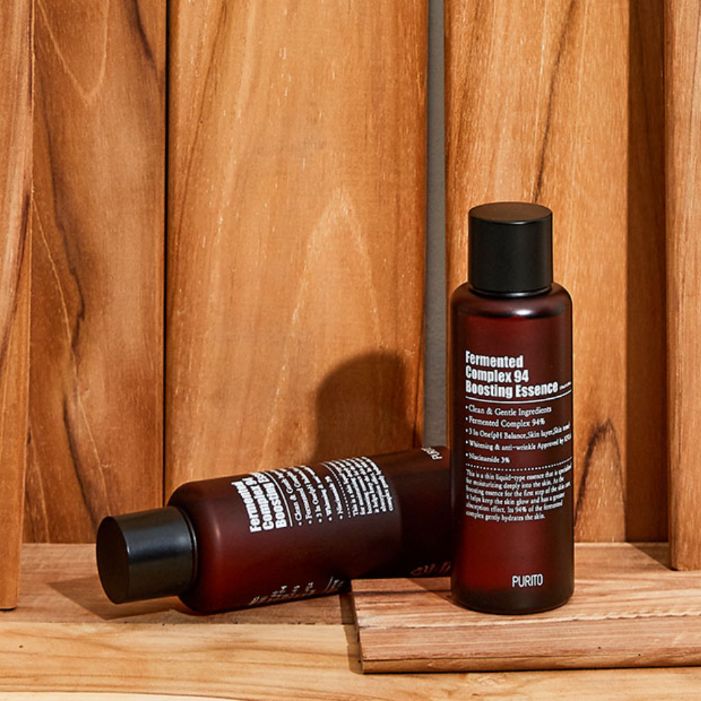Dve fľaše PURITO Fermented Complex 94 Boosting Essence 150ml na zníženie jemných liniek a vrások sediace na drevenom stole.