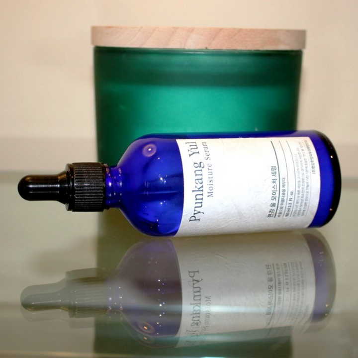 Fľaša Pyunkang Yul Moisture Serum 100ml vyrovnáva úroveň vlhkosti na stole vedľa sviečky.