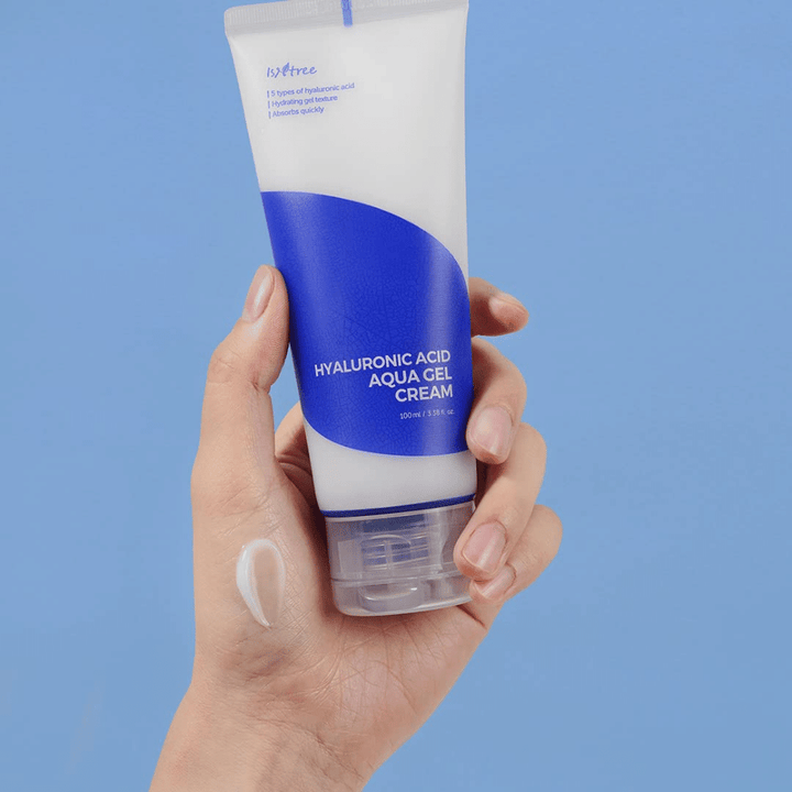 Ruka, ktorá drží tubu Isntree Hyaluronic Acid Aqua Gel Cream 100ml na modrom pozadí.